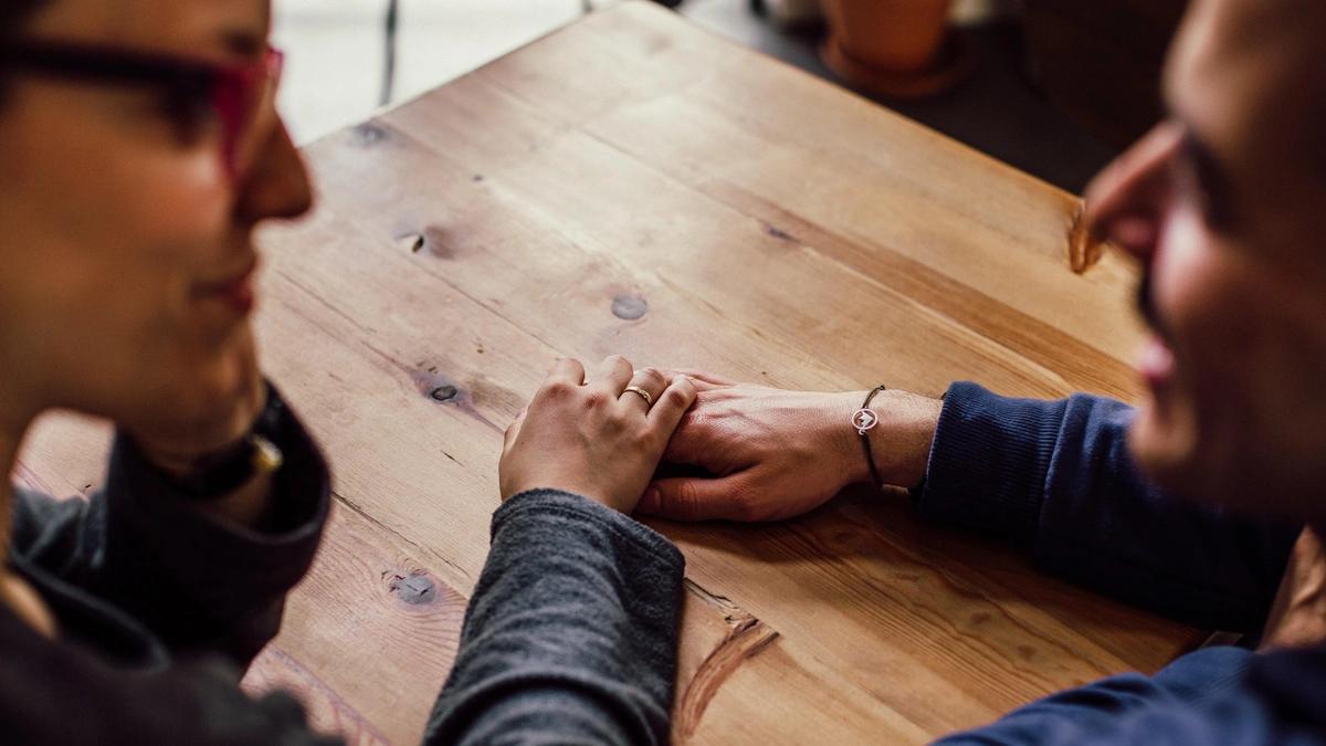 a couple sits at a wooden table with a hand on the table, one hand on top of the other. It is zoomed in and focused on their hands. Their faces are blurred.